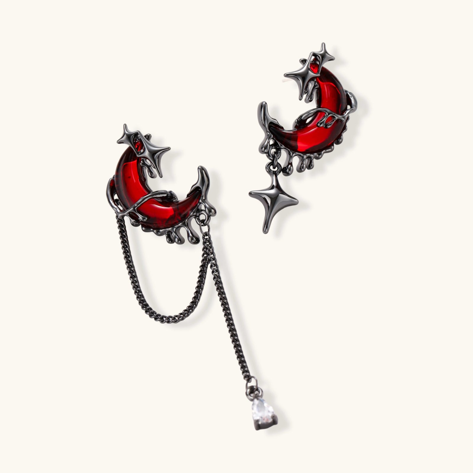 Gothic asymmetrical earrings with red crescent moons, black dripping stars, chains and crystal drop on white background.
