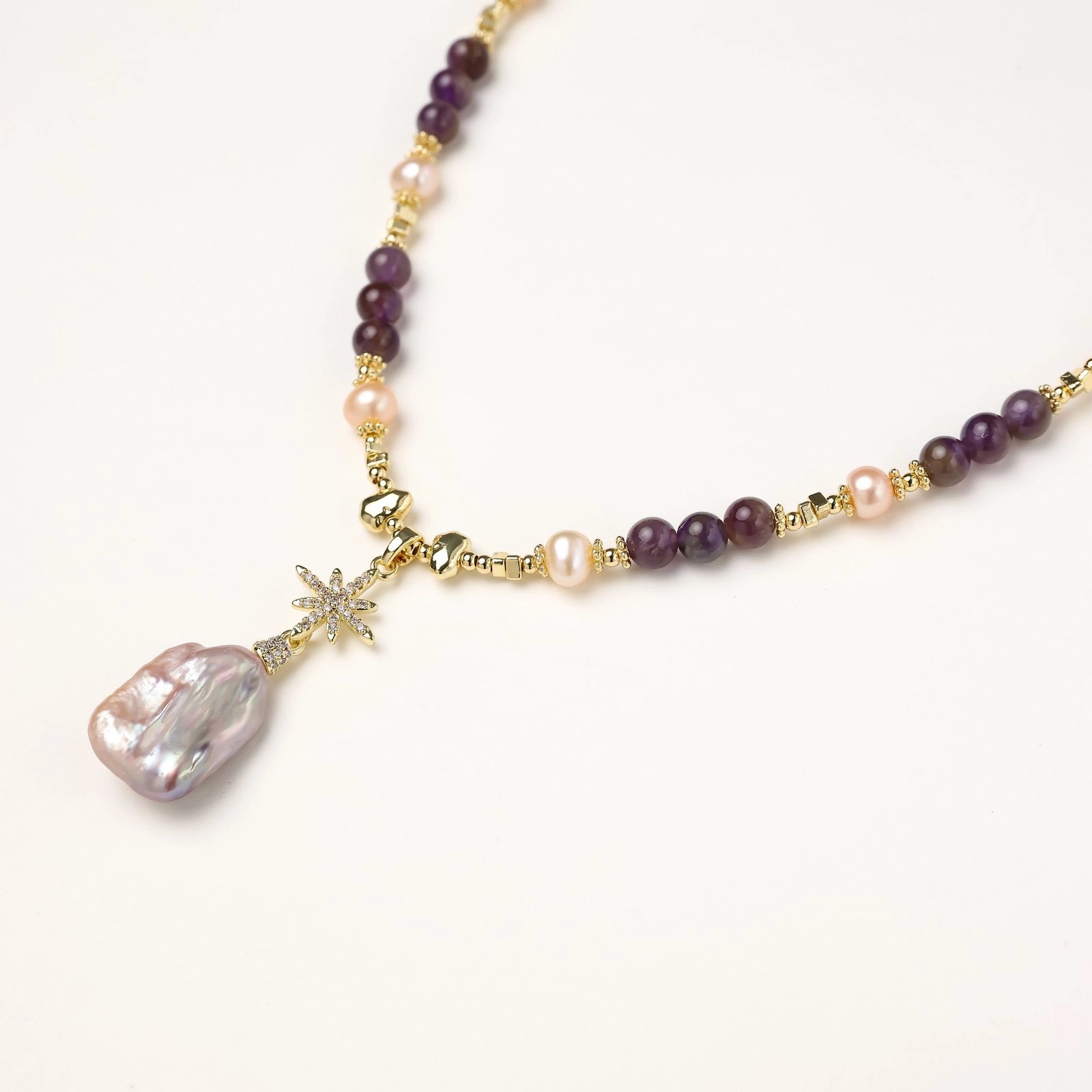 Gold necklace with iridescent baroque pearl pendant, purple amethyst beads, and crystal star charm on cream background