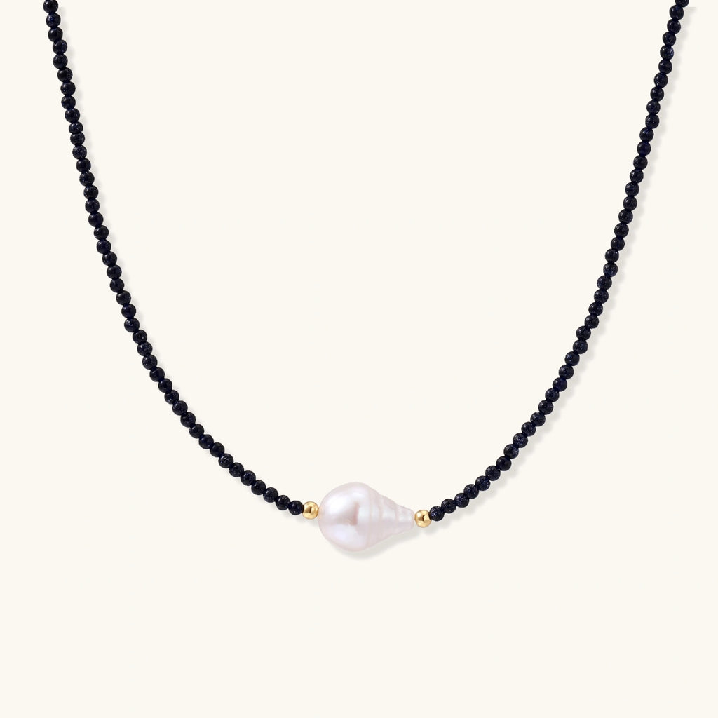 Delicate necklace with black blue sandstone beads and baroque pearl center on white background with gold accents.