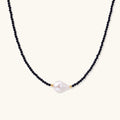 Delicate necklace with black blue sandstone beads and baroque pearl center on white background with gold accents.