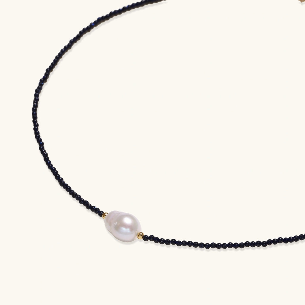 Delicate necklace with black blue sandstone beads and baroque pearl center on white background with gold accents.