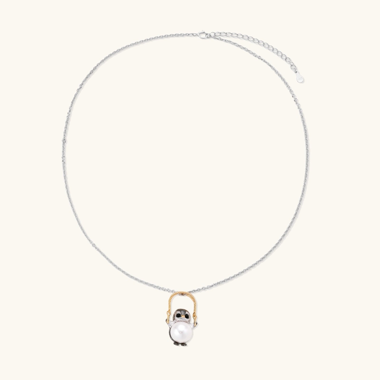 Whimsical owl pendant holding a white pearl on a gold swing, rhodium-plated silver chain on cream background.