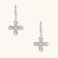 Ornate white gold-plated cross earrings with baguette-cut zirconia stones and twisted leverback hooks on beige background