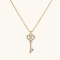 Delicate gold key pendant with heart-shaped bow and crystal pavé detailing on fine chain against cream background.